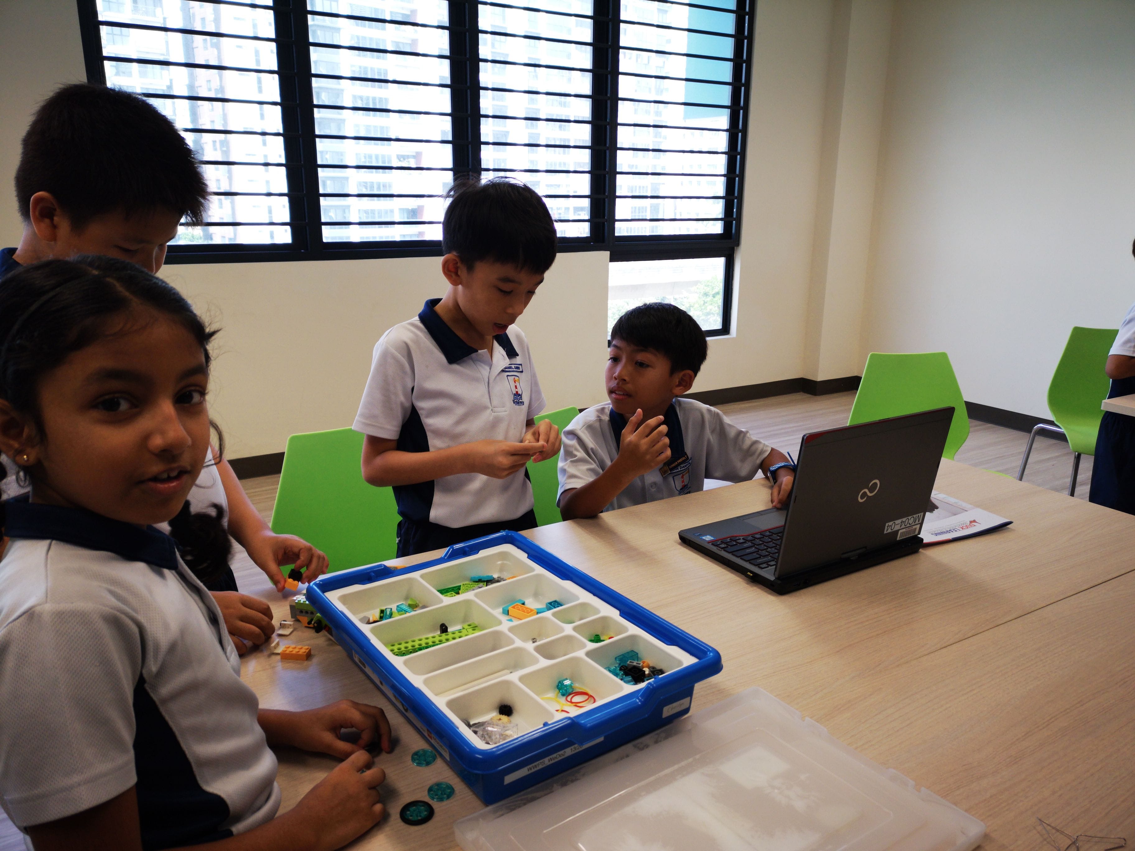 Four school children in uniforms work with a LEGO kit and a laptop at a classroom table.